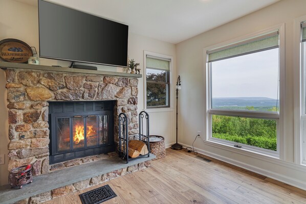 A stone fireplace with a TV above it, a large window with a view, and a basket of firewood.