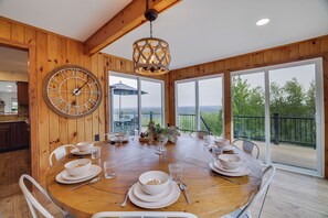 A wooden dining table set with white plates, bowls, and glasses, surrounded by white chairs, with a large wall clock and a hanging light fixture. - Fall Foliage Mountainside Views, Minutes To Jim Thorpe, Hot Tub & Game Room (Jim Thorpe)