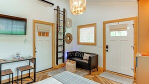 Ein modernes Wohnzimmer mit einem schwarzen Ledersofa, einem Holztisch und einer Deckenleuchte. An der Wand lehnt eine Leiter und es gibt eine Tür mit einem Fenster.