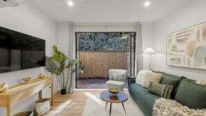 Een moderne woonkamer met een groene bank, een ronde salontafel, een aan de muur bevestigde televisie en een houten omheining die naar een tuin leidt.