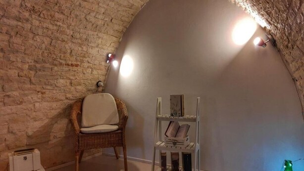 Une pièce chaleureuse avec un mur en briques courbé, un fauteuil en osier, une étagère blanche remplie de livres et une bouteille verte.