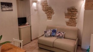 Un salon chaleureux avec un canapé beige, une petite table et un téléviseur. On y trouve des coussins décoratifs et un mur dont la texture rappelle celle de la pierre.
