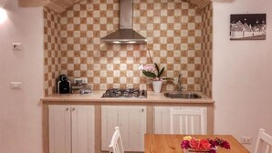 Une cuisine avec un crédence à carreaux, une hotte en acier inoxydable, une cuisinière à gaz et un évier. On y trouve un meuble blanc sous le plan de travail et une chaise devant une table en bois sur laquelle est posé un vase de fleurs.