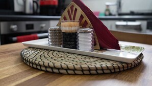 A kitchen counter with a woven placemat, a white tray with cocktail sticks, salt and pepper shakers, and a red napkin holder.