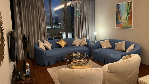 A modern living room with a blue sectional sofa, a white armchair, and a glass coffee table. There is a large window with curtains, a painting on the wall, and a chandelier. - Luxury Stay (6th of October City)
