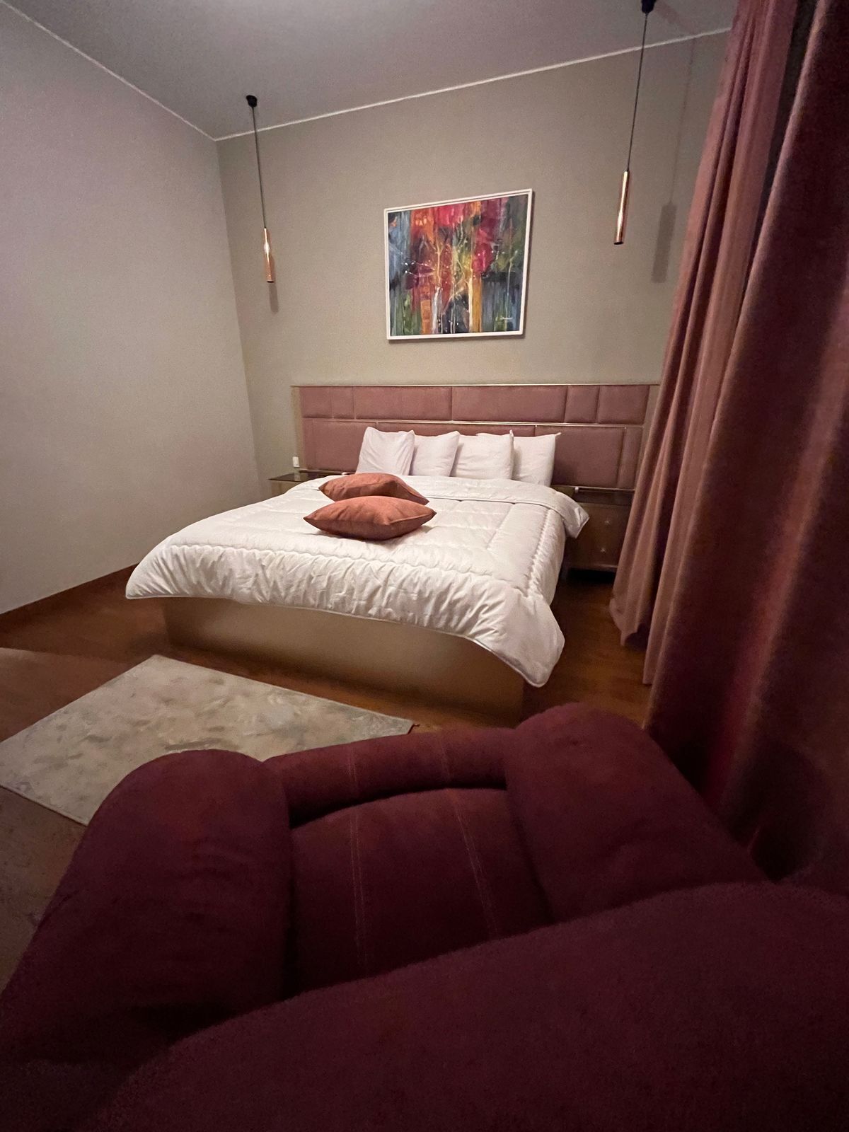 A bedroom with a bed, a headboard, a painting on the wall, and a window with curtains.