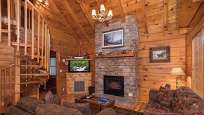 A log cabin interior with a stone fireplace, wooden staircase, and a cozy seating area. - Unforgettable Luxury Mountain Getaway (PIGEON FORGE)