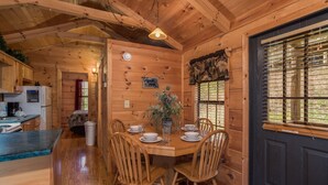 A wooden cabin interior with a dining table set for four, wooden chairs, a kitchen area with a refrigerator, and a window with a view of the outdoors.
