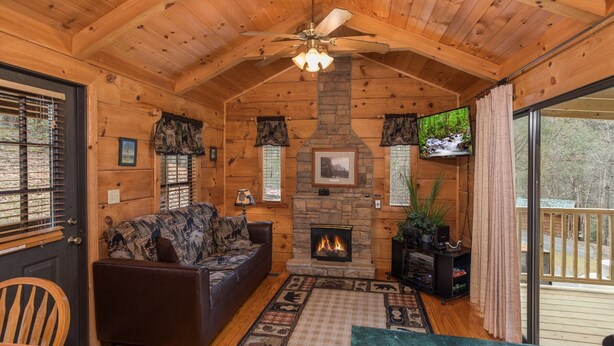 A cozy cabin interior with a fireplace, wooden walls, a ceiling fan, and a large window overlooking a deck.