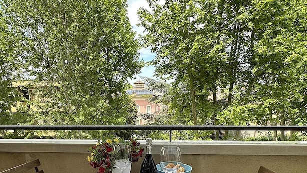 Una mesa en el balcón con una botella de vino, copas y un cuenco de aperitivos, rodeada de frondosos árboles verdes.