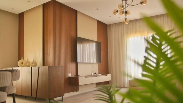 A modern living room with a flat-screen TV mounted on the wall, a wooden cabinet, and a large window with curtains. - Settle Inn Cairo Festival City Villa (New Cairo)