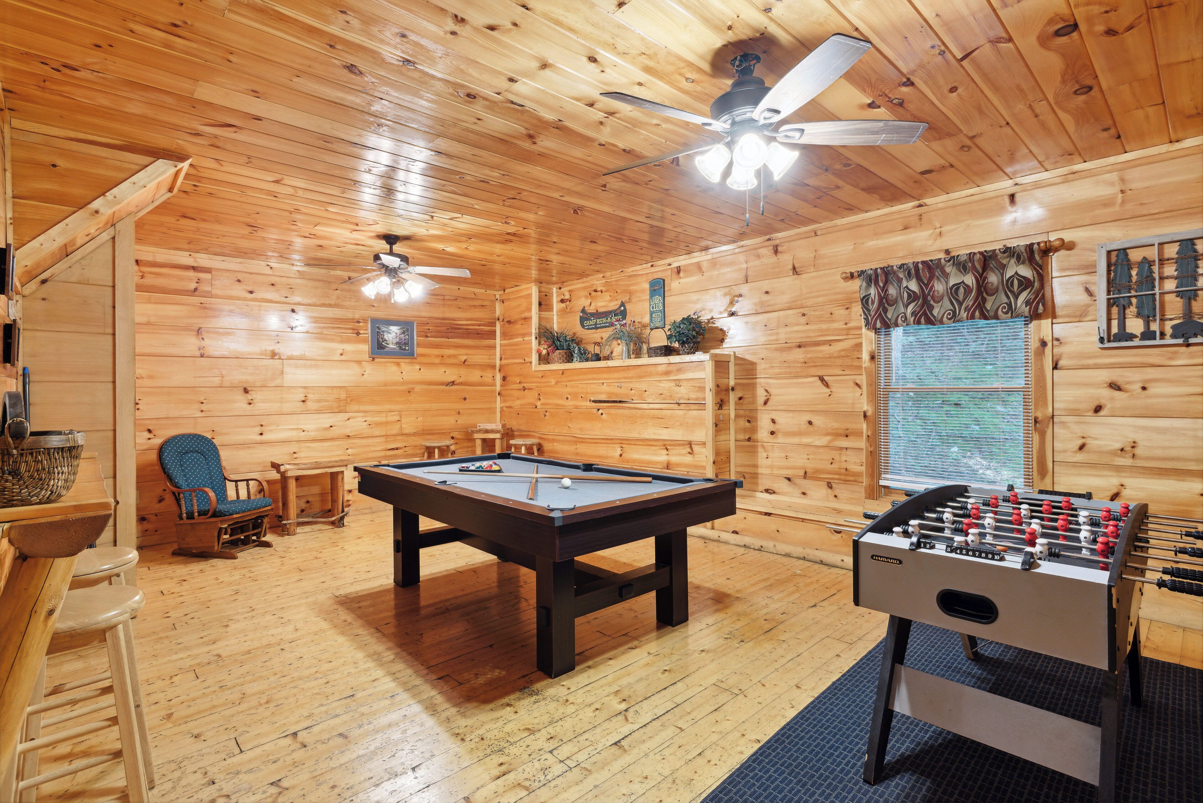 A wooden-floored room with a pool table, a foosball table, and a ceiling fan.