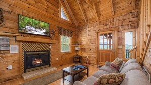 A cozy cabin interior with a fireplace, a television mounted above it, a sofa with decorative pillows, and a window overlooking a forested area. - Peace And Quiet With Incredible Views (PIGEON FORGE)
