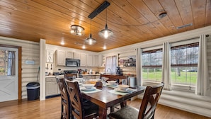 A wooden-paneled kitchen with white cabinets, a dining table set for four, and a view of the outdoors through large windows. - Creekside Haven equals a Heavenly Escape (PIGEON FORGE)