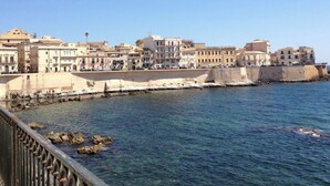 A coastal cityscape with buildings along the waterfront, a clear sky, and rocky outcrops in the water. - Mandolino Apartment - Two Bedroom Apartment, Sleeps 5 (Syracuse)