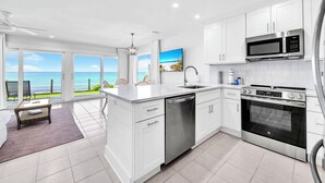Eine moderne Küche mit weißen Schränken, einem Edelstahl-Geschirrspüler und einem Backofen. Sie verfügt über eine große Kochinsel mit Spüle und Meerblick durch große Fenster.