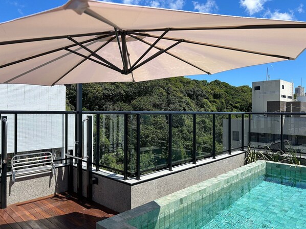 Una piscina en la azotea con barandilla de vidrio, un parasol color beige y vistas a un área boscosa.