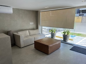 A modern living room with a sofa, a brown ottoman, and two potted plants. - Bela Hospedagem -  303 Bauten Aurea  (João Pessoa)