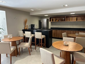 A modern kitchen with a central island, bar stools, and a dining area with round tables. - Bela Hospedagem -  303 Bauten Aurea  (João Pessoa)