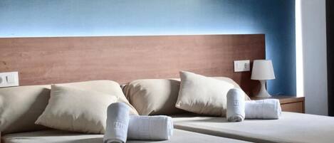 A hotel room with two beds, beige bedding, a wooden headboard, a wall-mounted lamp, and a window with curtains.