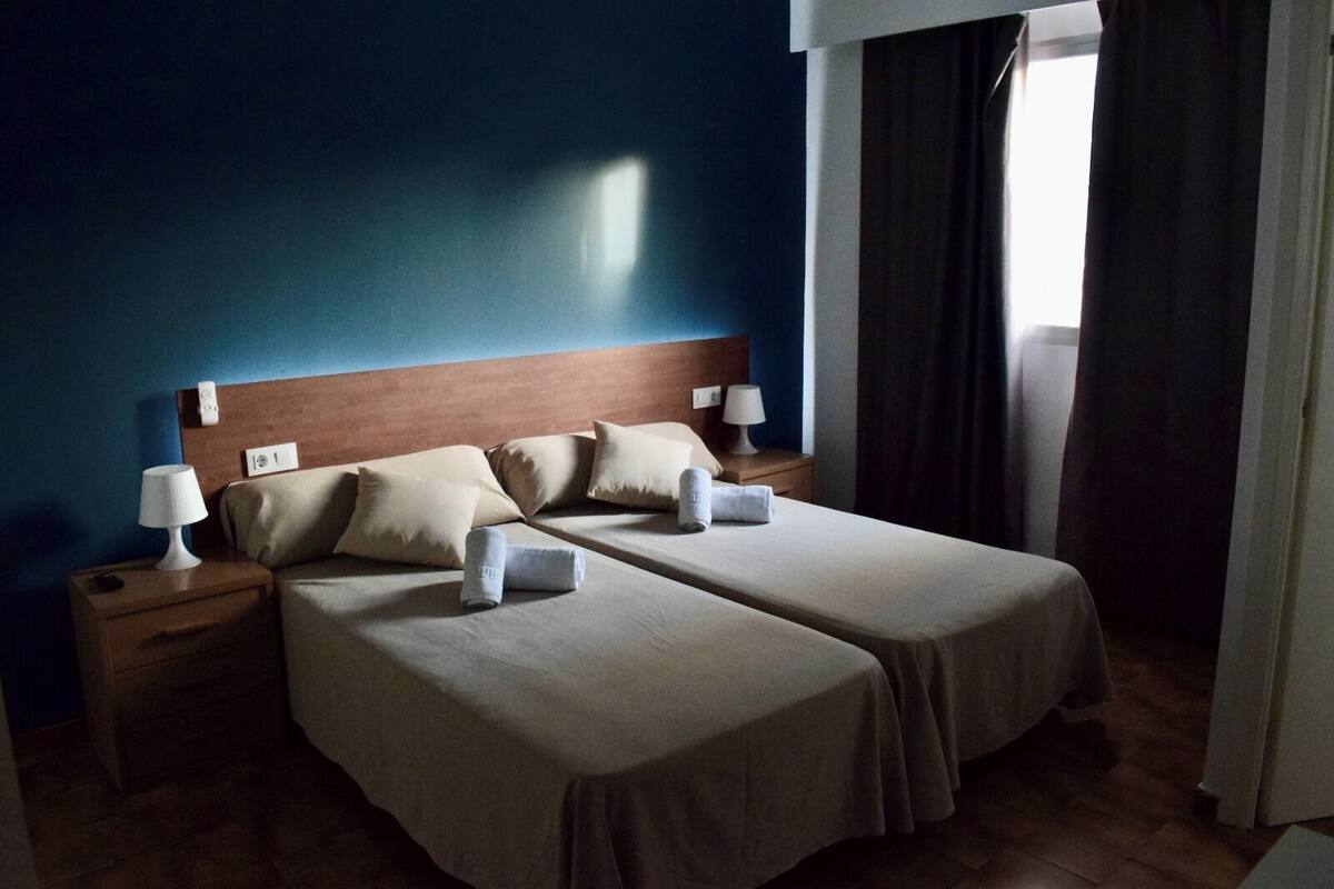 Ein Doppelbett mit zwei Kissen, ein Nachttisch mit Lampe und ein Fenster mit Vorhängen.