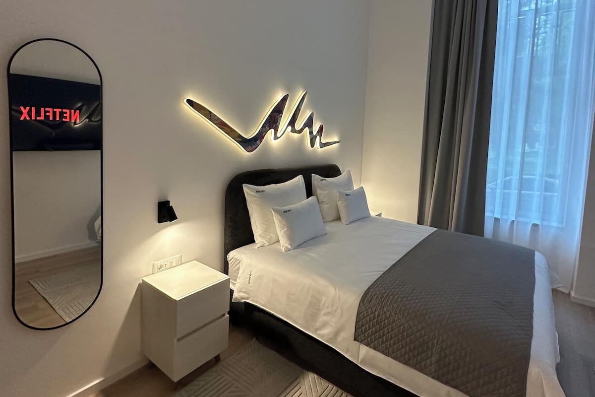A modern bedroom with a large bed, a bedside table, a wall-mounted mirror, and a decorative light fixture.