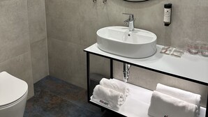 A modern bathroom with a round mirror, a white sink, and a toilet. There are towels and toiletries on the sink counter. - Hotel Eleven (Sarajevo)