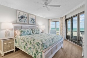 Un dormitorio con una cama grande, dos mesitas de noche, un ventilador de techo y vista a la playa a través de un ventanal corredero.