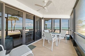 Un espacio al aire libre cubierto con ventilador de techo, puertas de vidrio que dan a vista de la playa y mobiliario exterior blanco.