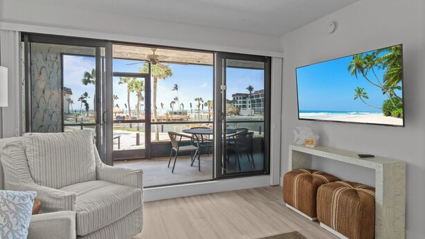Ein Zimmer mit einer großen Schiebeglasdoor zum Balkon mit Blick auf Strand und Palmen. An der Wand ist ein Fernseher angebracht und es gibt ein Sofa mit einem Konsolentisch.