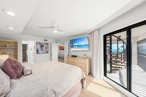 A bedroom with a large bed, a dresser, a television, and a balcony with a view of the beach.