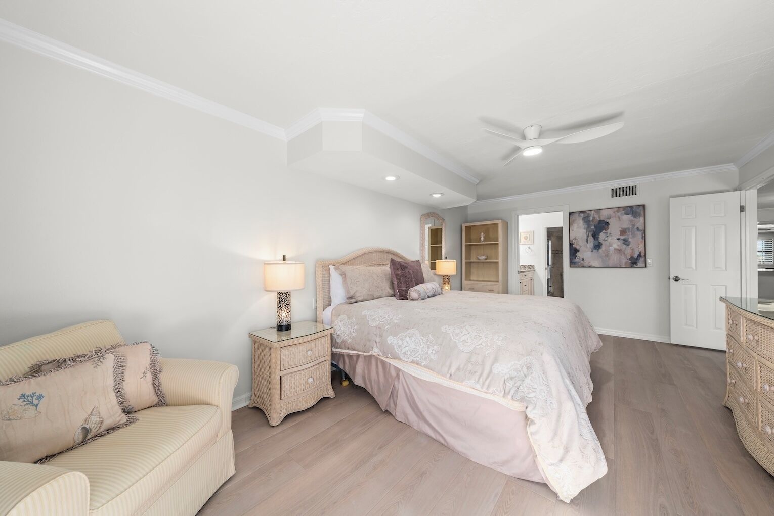A bedroom with a large bed, two bedside tables, a nightstand, a ceiling fan, and a door leading to another room.