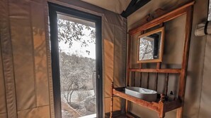 A bathroom with a wooden vanity, a sink, and a mirror. There is a window with a view of trees and a building outside. - Timbila Game Lodge (Phalaborwa)