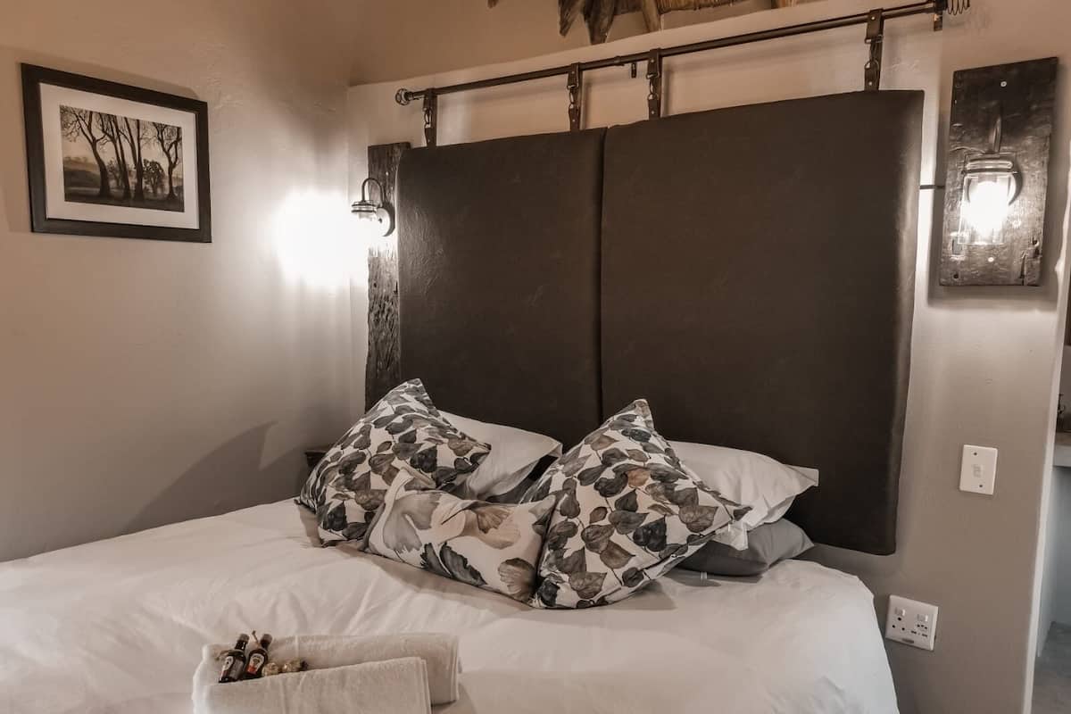 A bed with a dark headboard, two pillows with a leaf pattern, a white blanket, and a small towel with bottles.