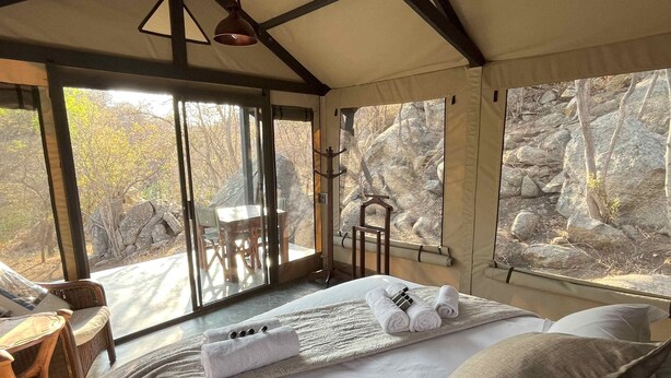A bedroom with a large bed, a wooden chair, a table, and a view of rocky terrain through the windows. - Timbila Game Lodge (Phalaborwa)