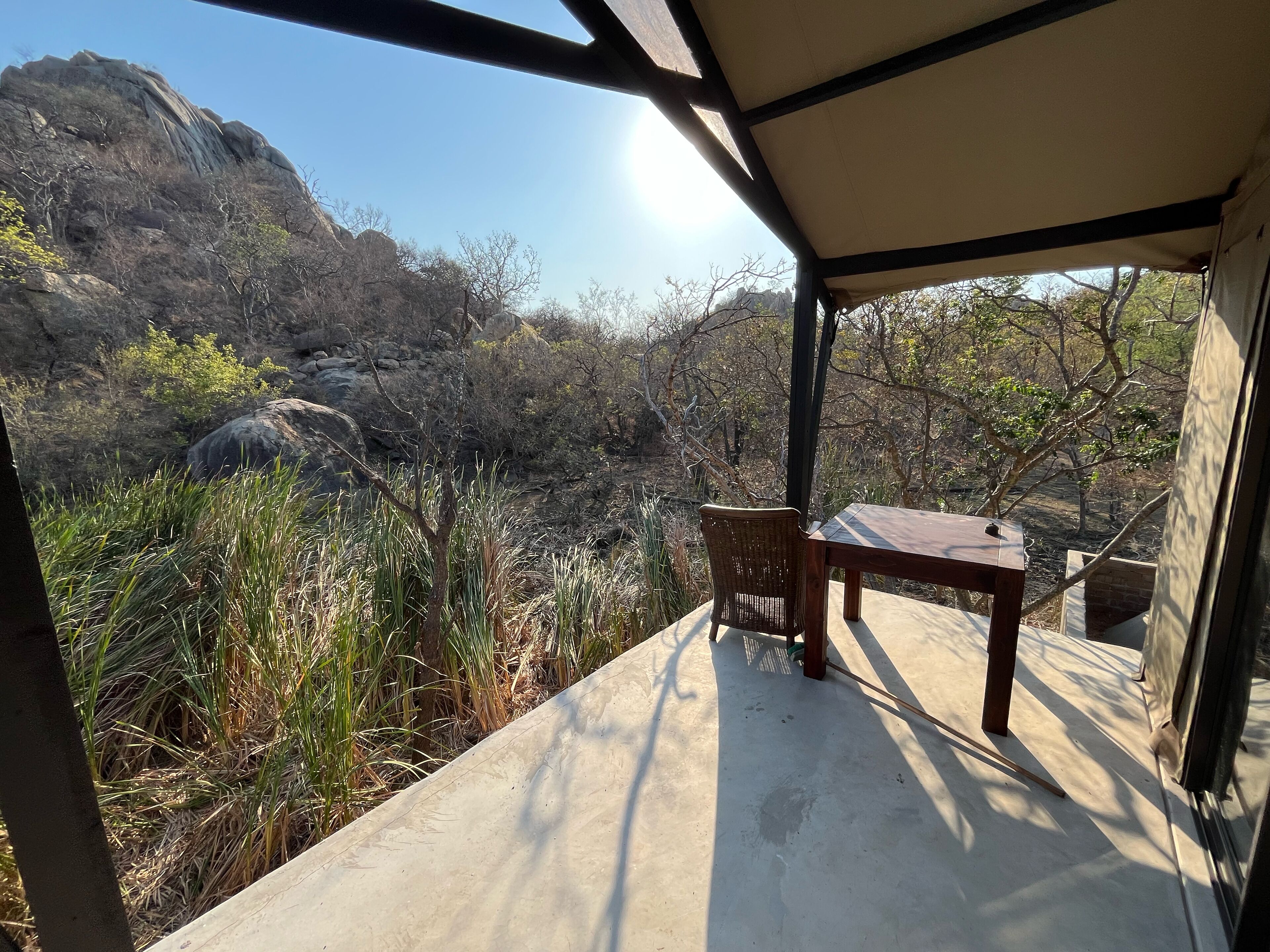 A covered outdoor seating area with a table and chair, overlooking a rocky landscape with sparse vegetation.