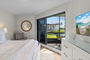A bedroom with a bed, a chair, a dresser, and a large window with a view of a beach and buildings.