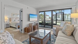 A modern hotel room with a bed, a sitting area with a glass table, and a balcony with a view of the beach and other buildings.