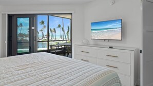 A bedroom with a large bed, a TV, and a view of a beach through a sliding glass door.