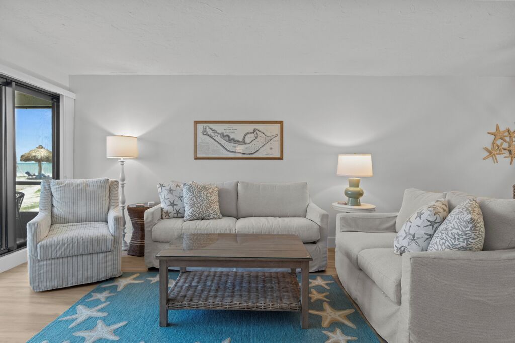 A living room with a sofa, armchair, and coffee table, a framed map on the wall, and a view of the sea through a large window.