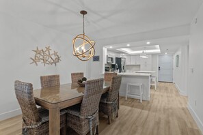 Un comedor moderno con una mesa de madera y sillas de mimbre, una lámpara colgante y una cocina al fondo.