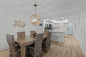 Un espace repas moderne avec une table en bois et des chaises en osier, un luminaire suspendu et une cuisine en arrière-plan.