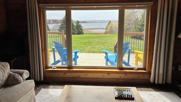 Une pièce chaleureuse avec une grande fenêtre donnant sur une terrasse où se trouvent des chaises bleues, et d’où l’on peut admirer un lac.