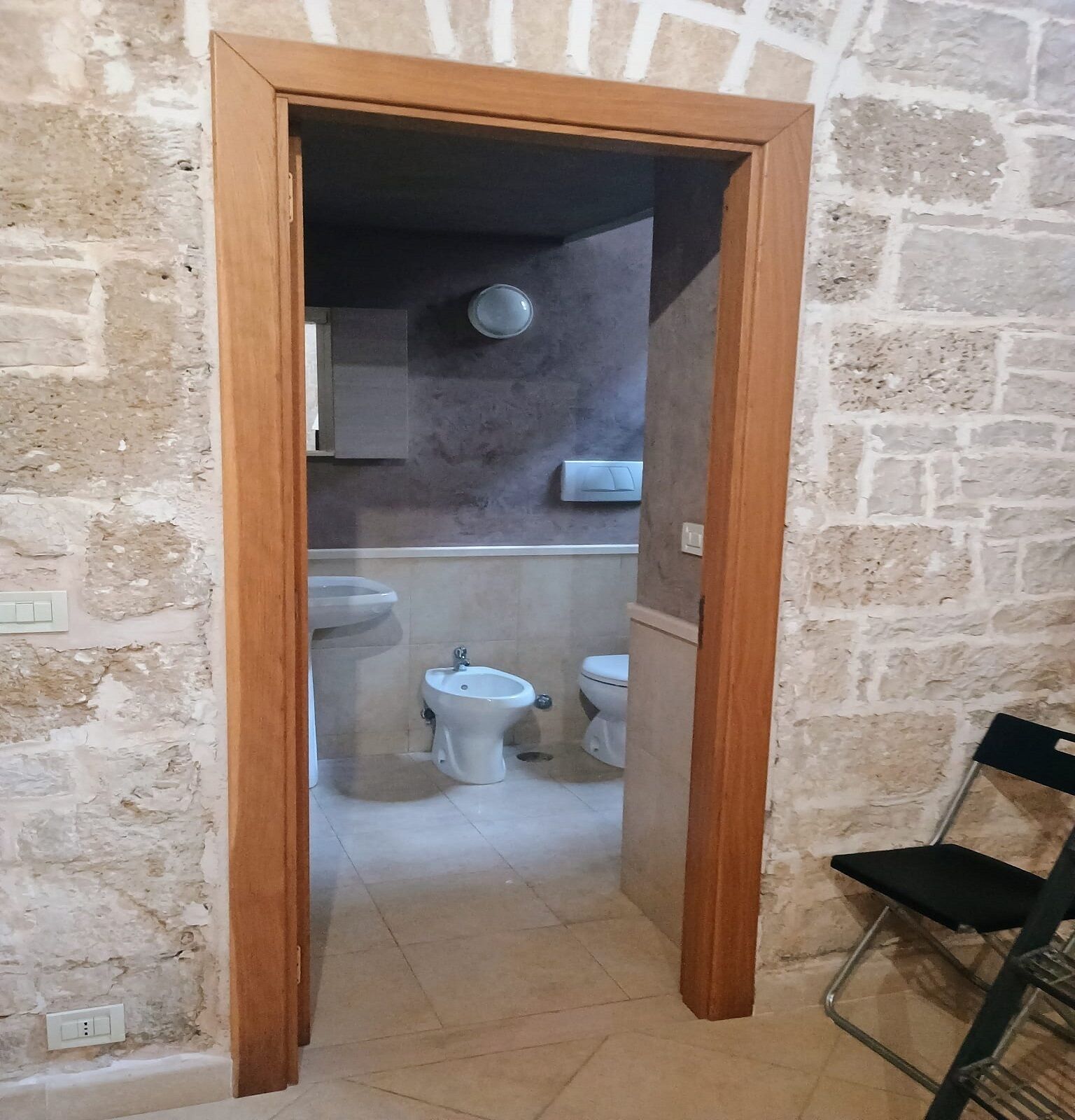 Ein schmaler Flur führt zu einem Badezimmer mit Waschbecken, Bidet und Toilette. Die Wände sind aus Stein, und der Türrahmen ist aus Holz.