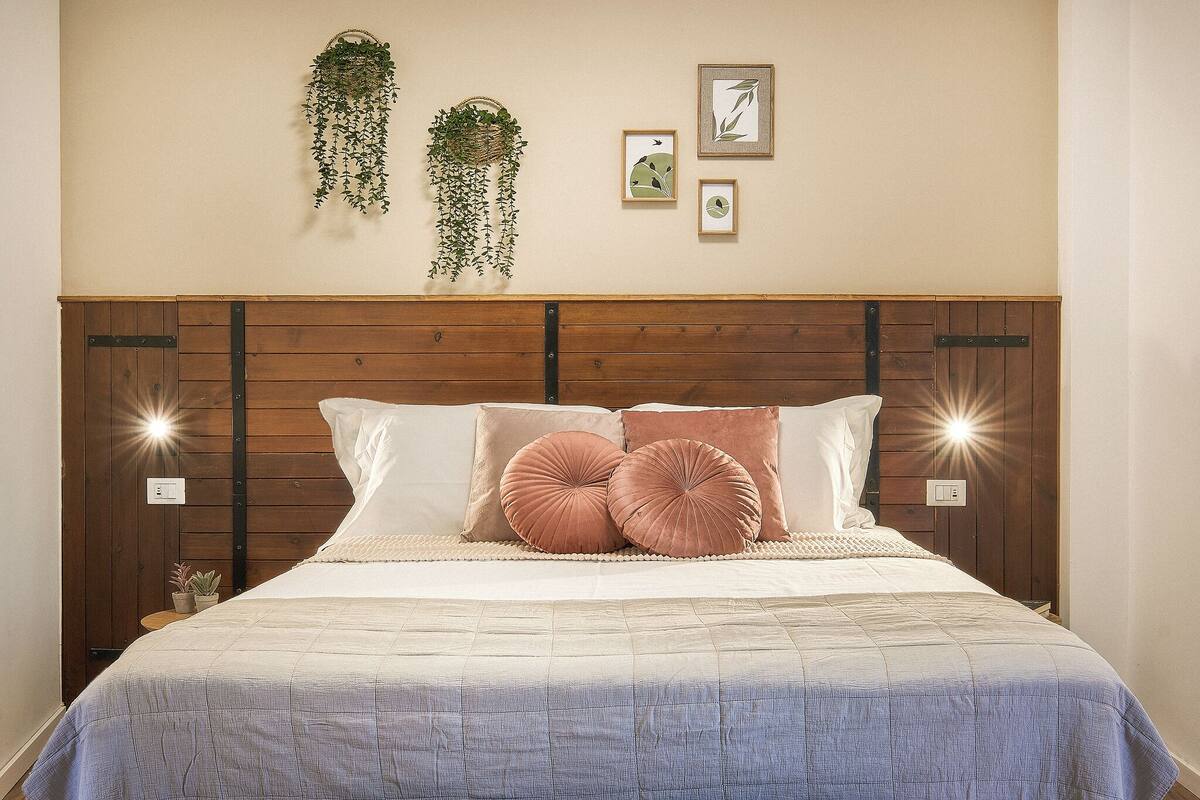 Ein ordentlich bezogenes Bett mit Holzkopfende, zwei großen Kissen und einer hängenden Pflanze an der Wand.