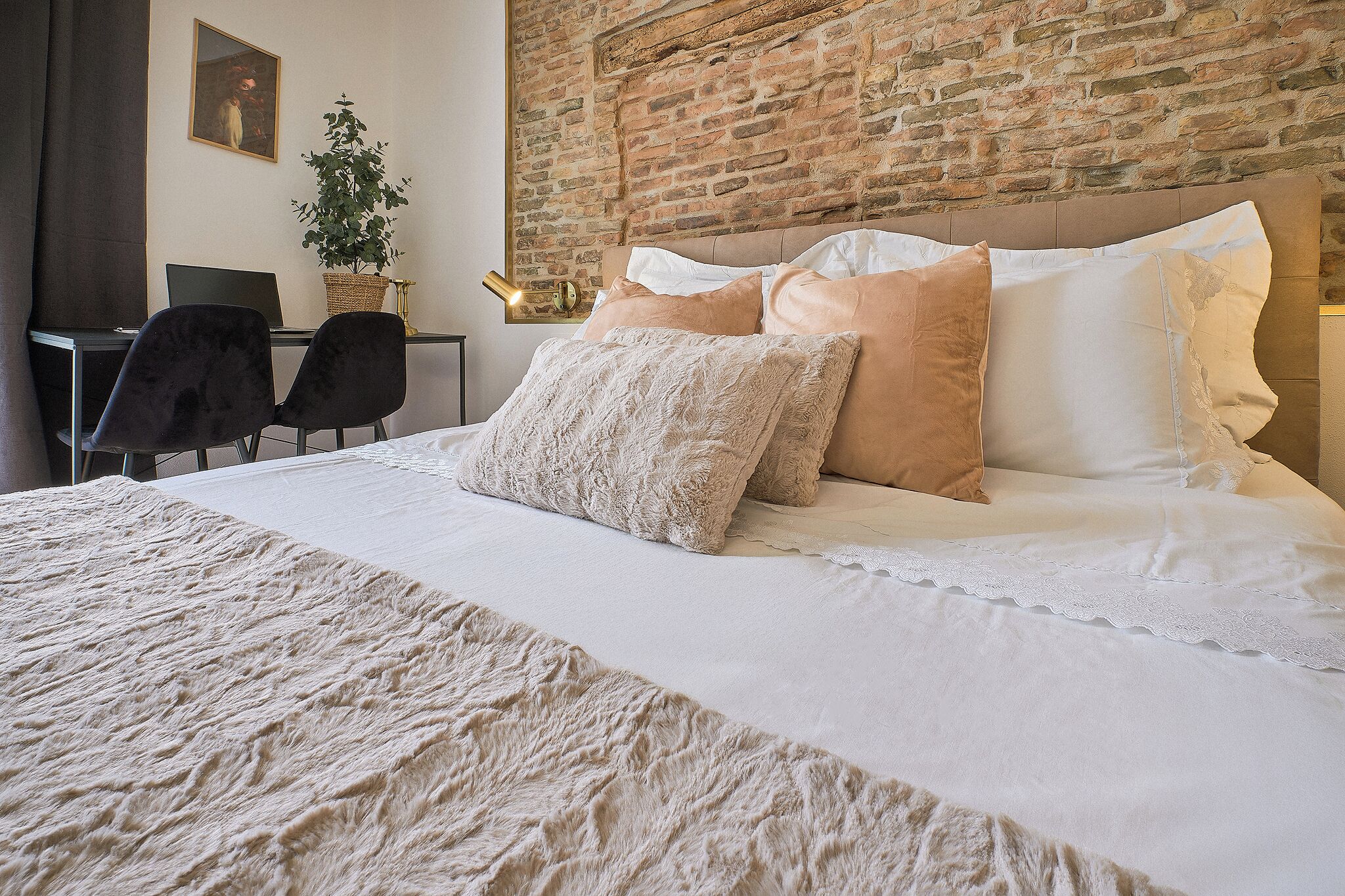 Un lit bien fait, avec une couette blanche et beige, un grand plaid beige et un mur en briques en arrière-plan.