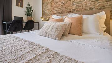 Un lit bien fait, avec une couette blanche et beige, un grand plaid beige et un mur en briques en arrière-plan.
