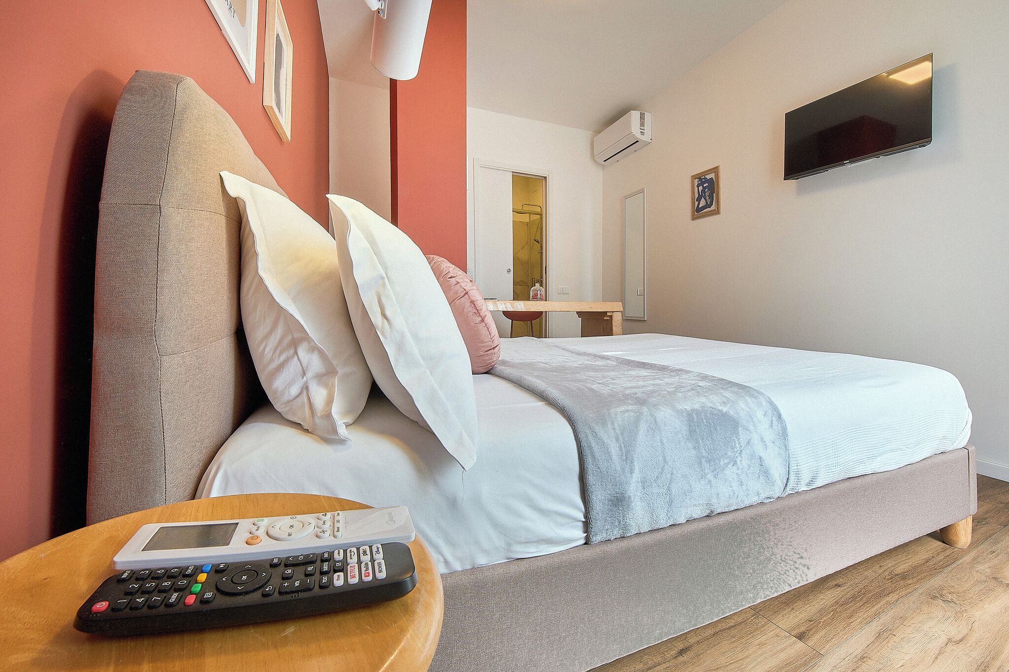 A bedroom with a bed, a bedside table with a remote control, a wall-mounted TV, and an air conditioning unit.