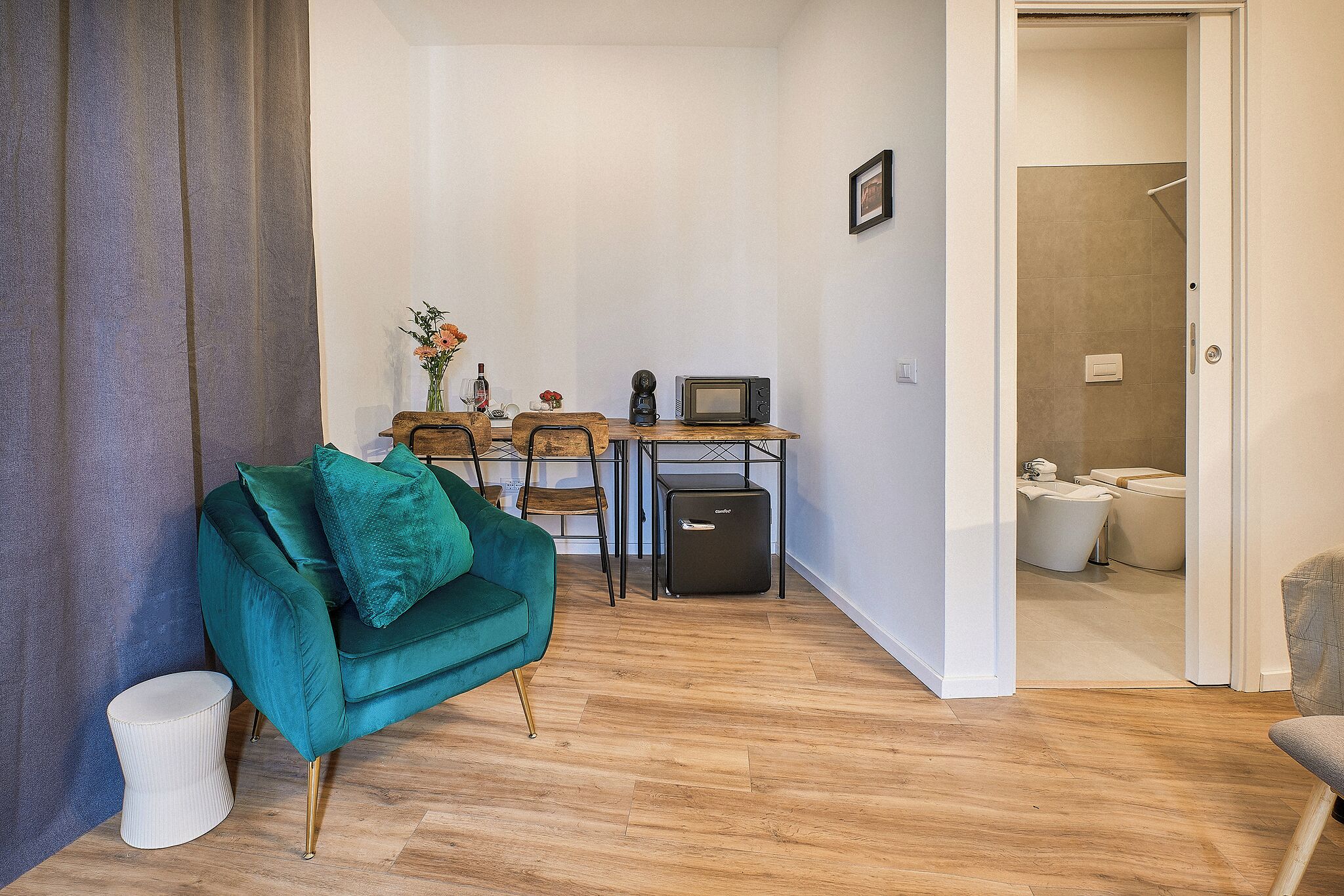 En kompakt opholdsstue med en turkis sofa, et lille træbord med mikroovn, og udsigt til et badeværelse med brusebad og toilet.