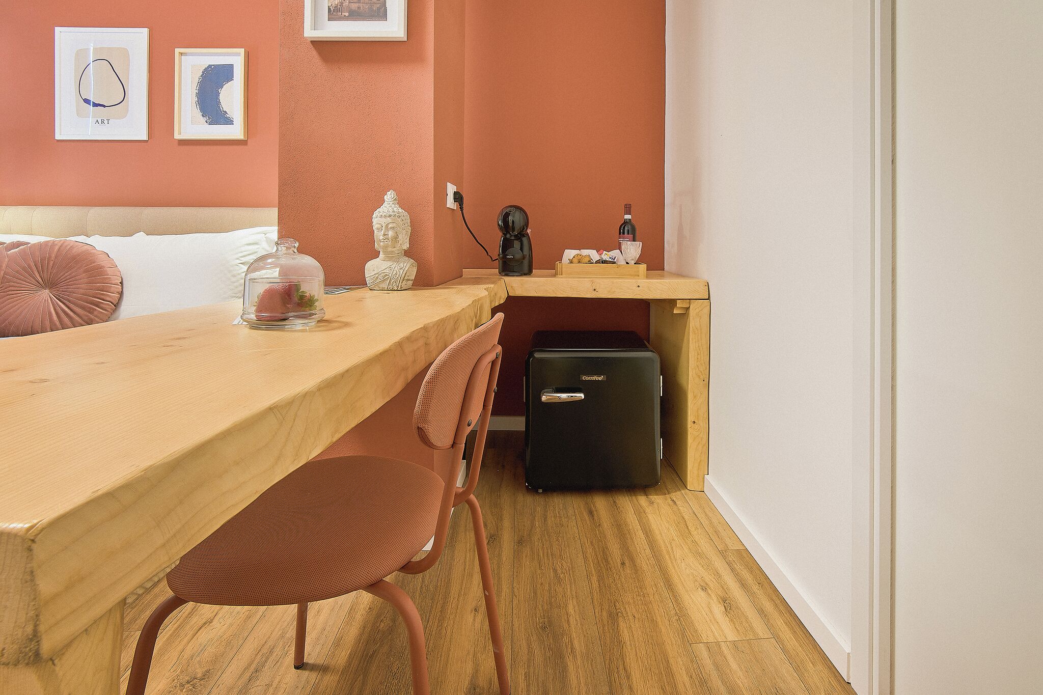 Una stanza con un tavolo di legno, una sedia, un letto con biancheria bianca, una valigia nera e una parete con due quadri.